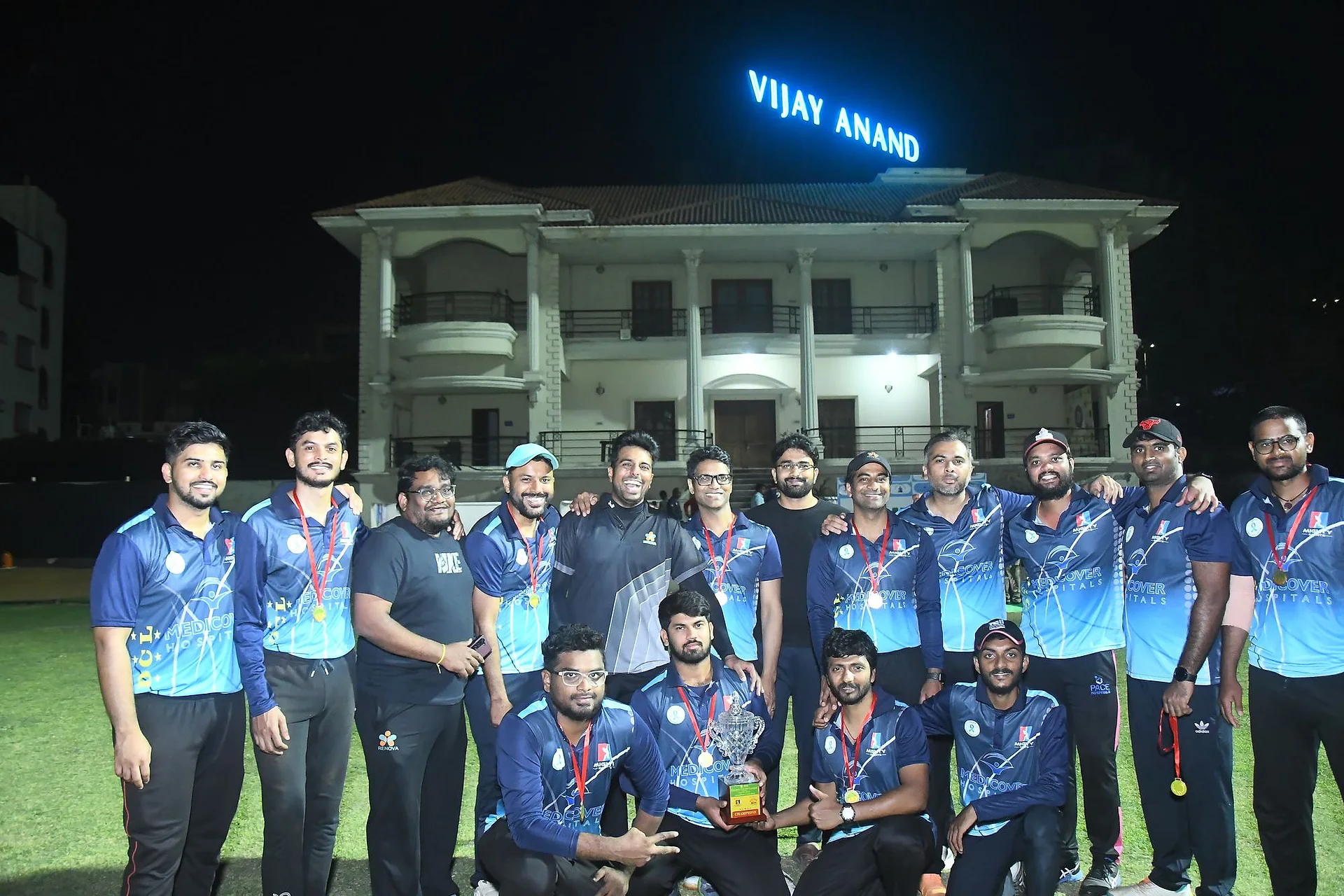 A cricket team of doctors in blue jerseys posing together on the ground at night