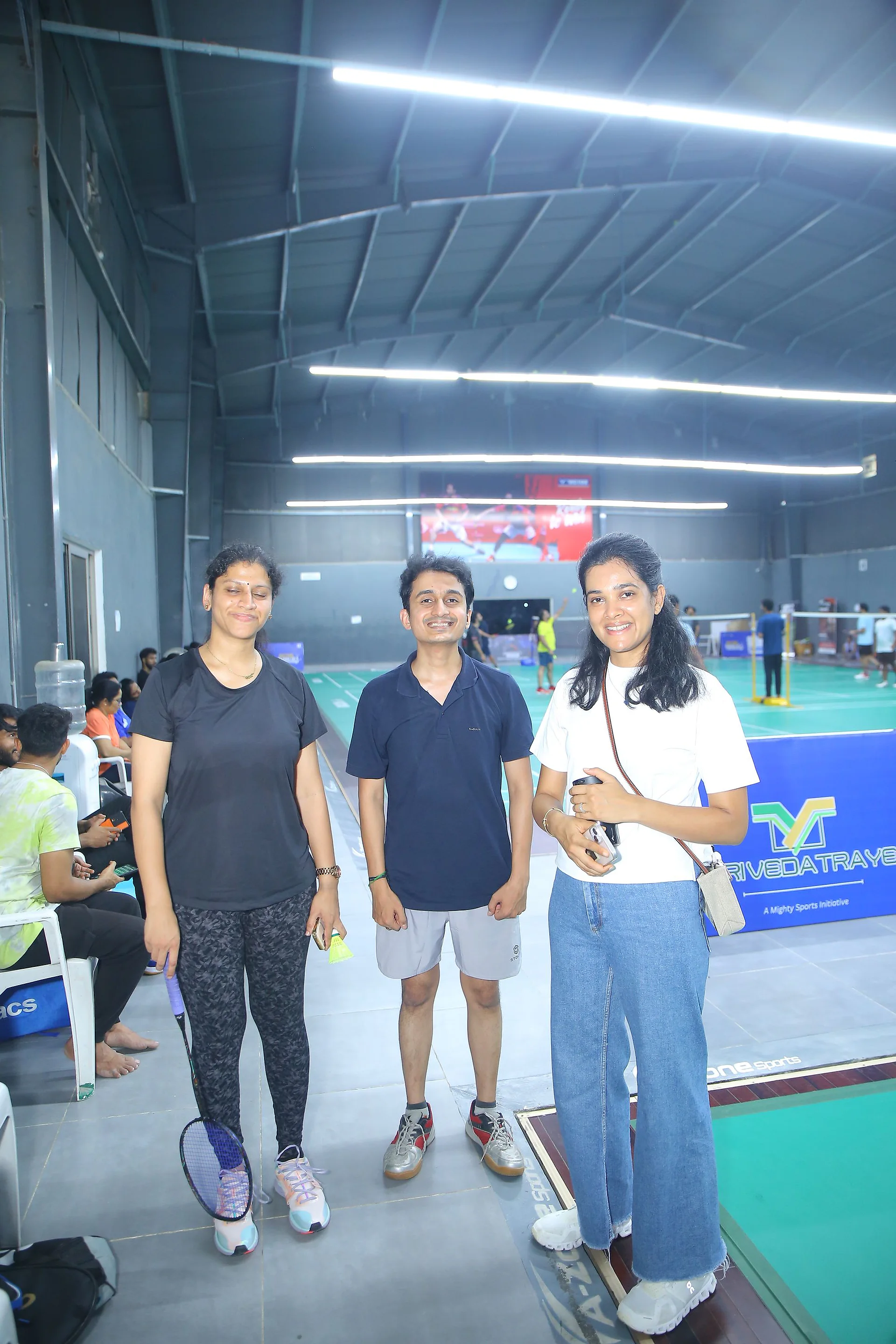 Three participants standing in the badminton hall with matches under way behind them