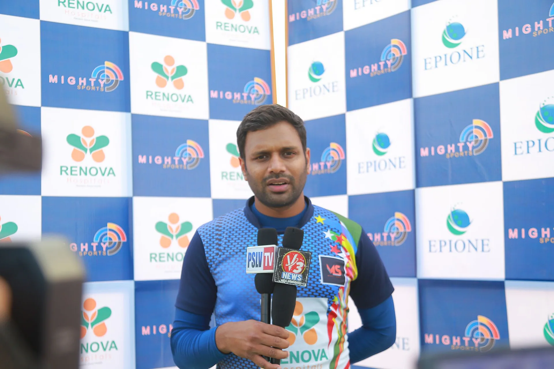 A broadcaster speaking into a microphone in front of the Mighty Sports backdrop