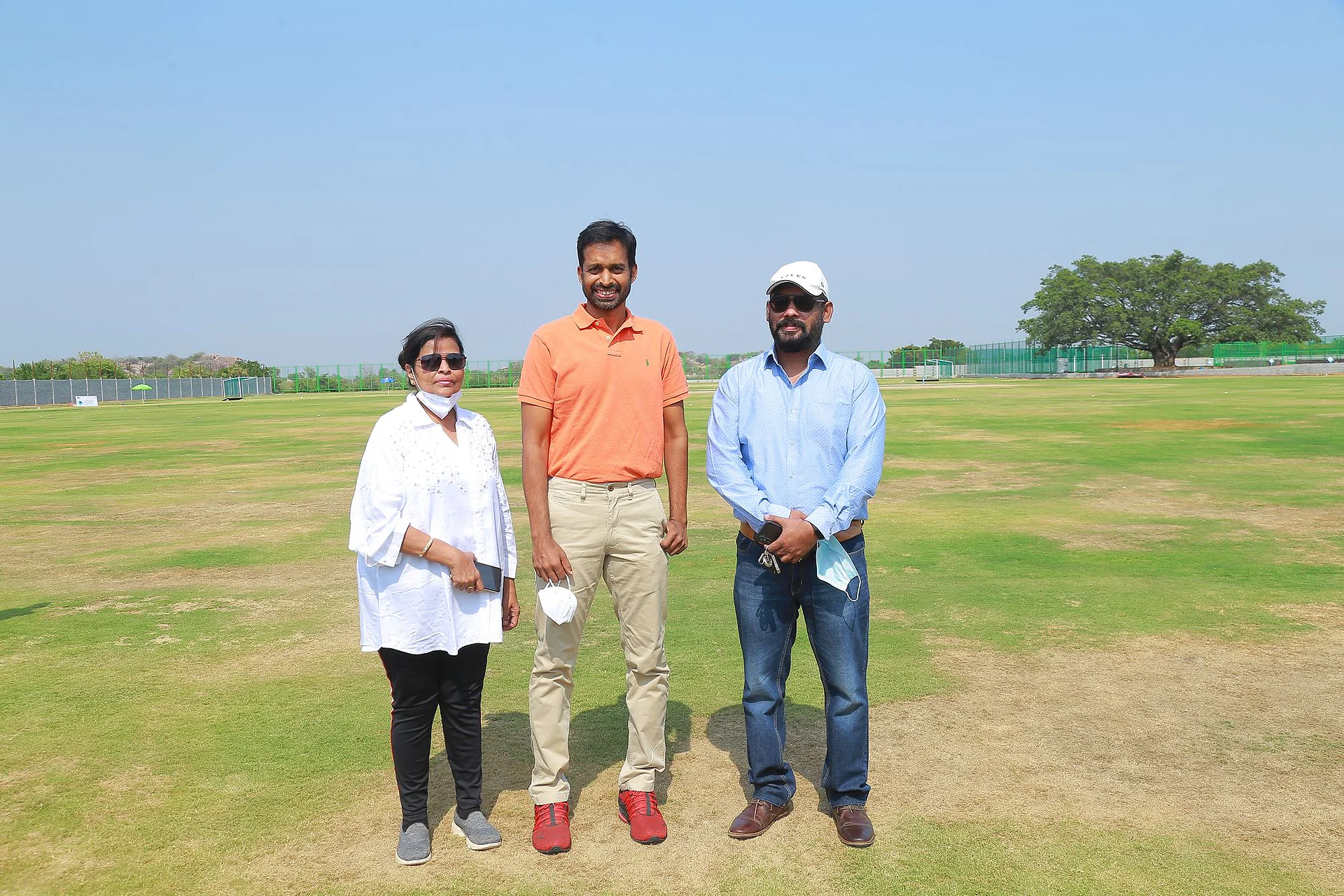 Three guests in casual clothes posing together on the open cricket ground under a clear blue sky