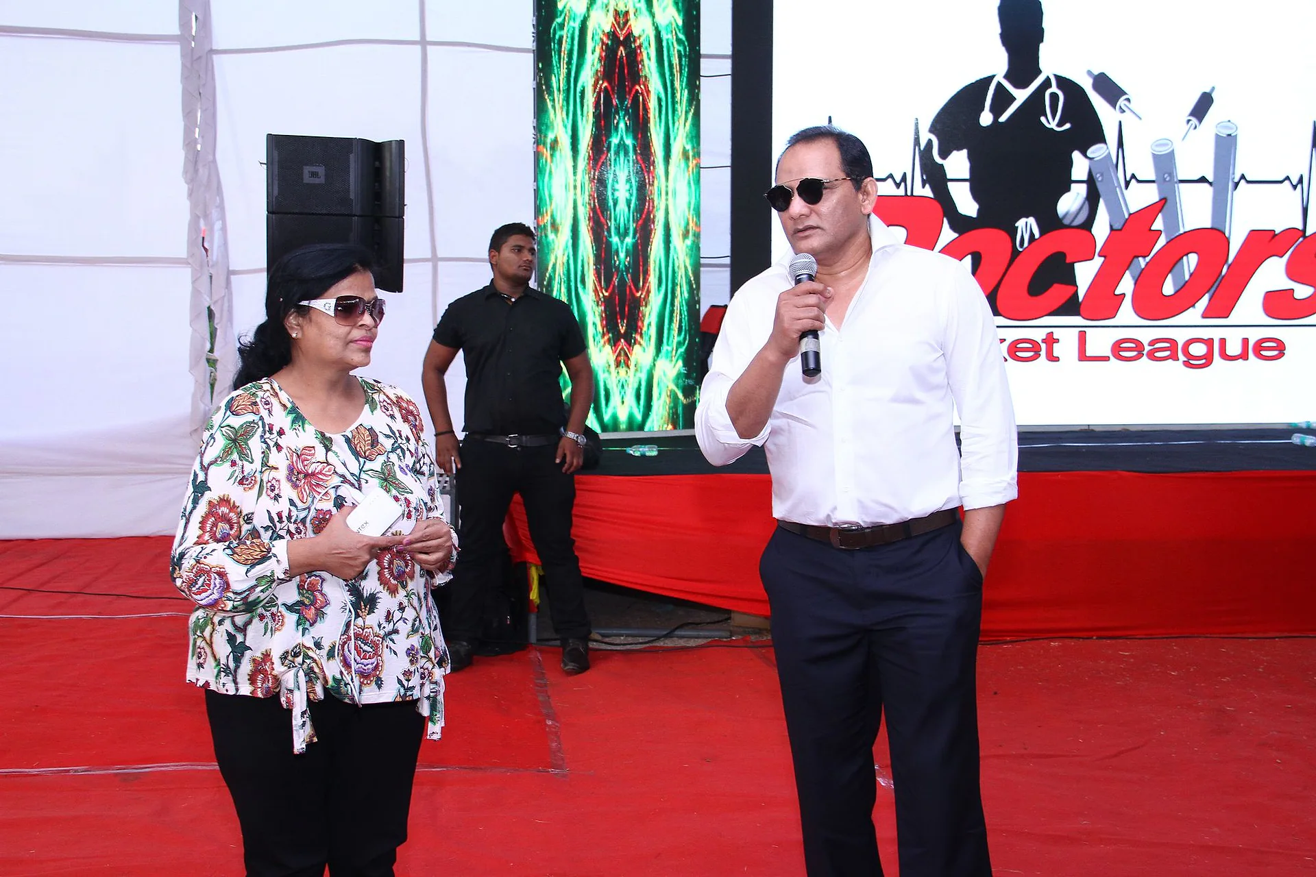 A guest in sunglasses speaking into a microphone beside a woman on the red-carpeted stage in front of the Doctors Cricket League screen