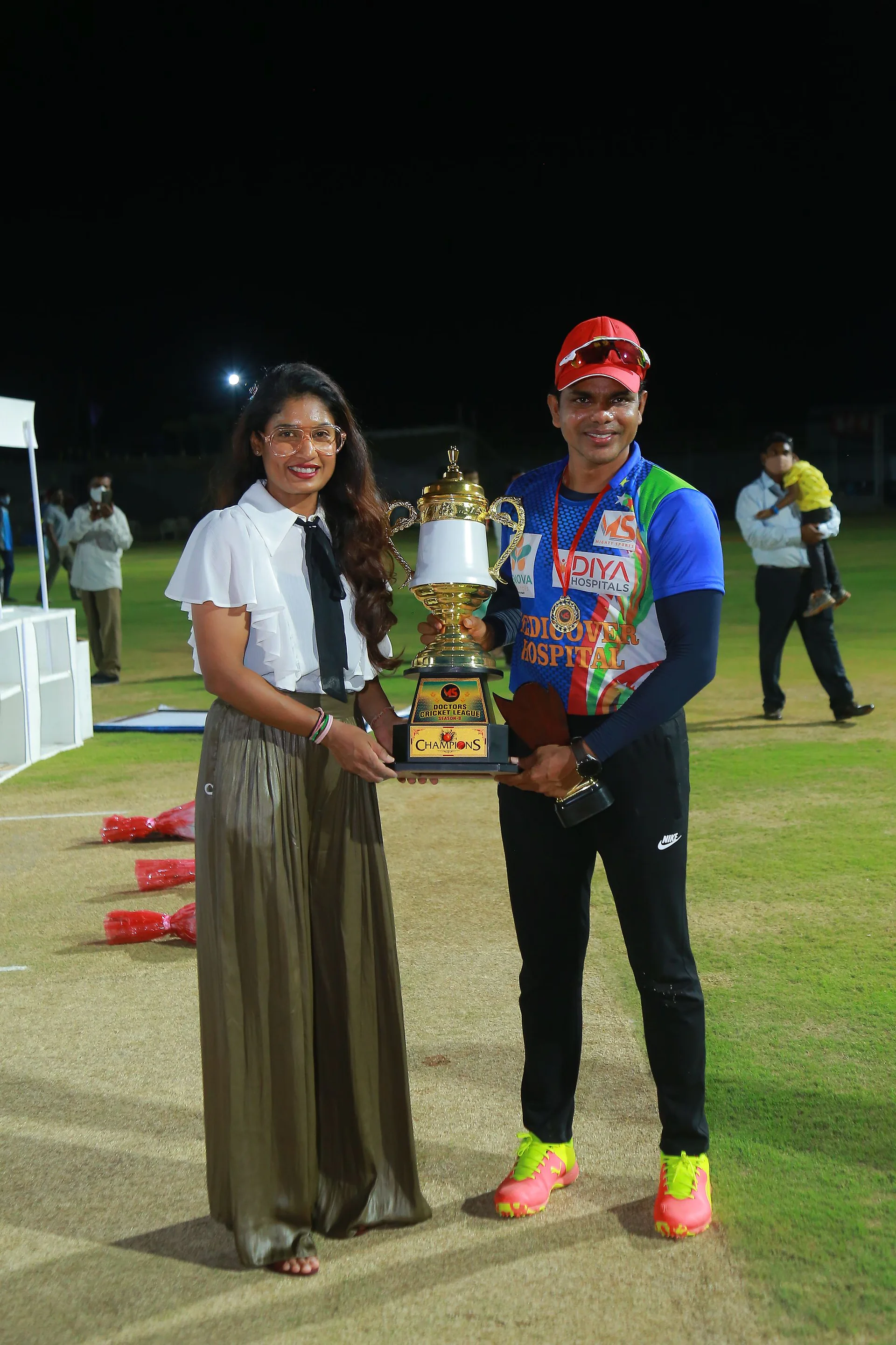 A guest in a white blouse presenting the Doctors Cricket League champions' trophy to a player in a blue team jersey and medal at night